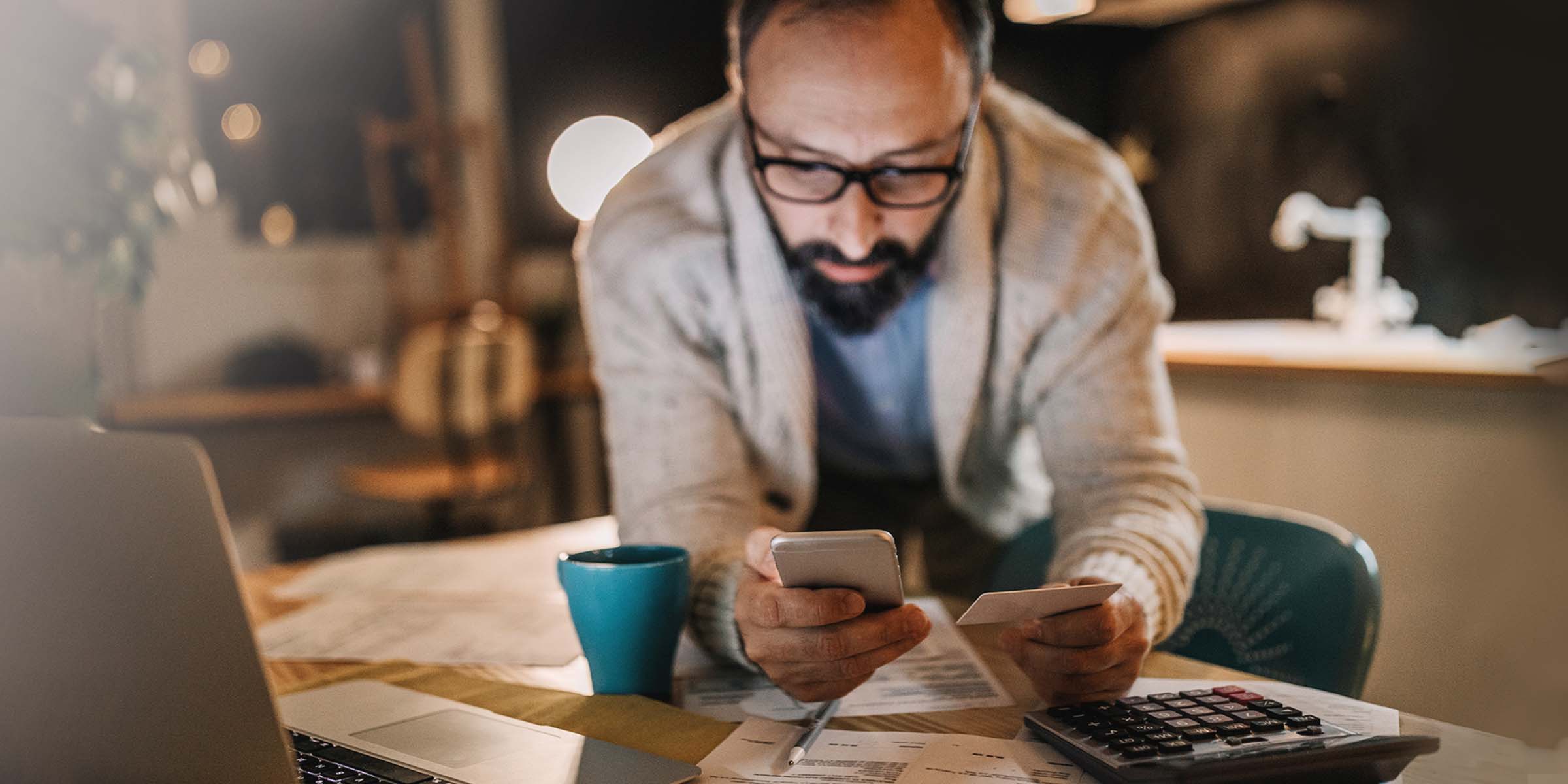 Tummahiuksinen parrakas mies syöttää pankkikortin numeroa kännykkään työpöytäänsä nojaten.