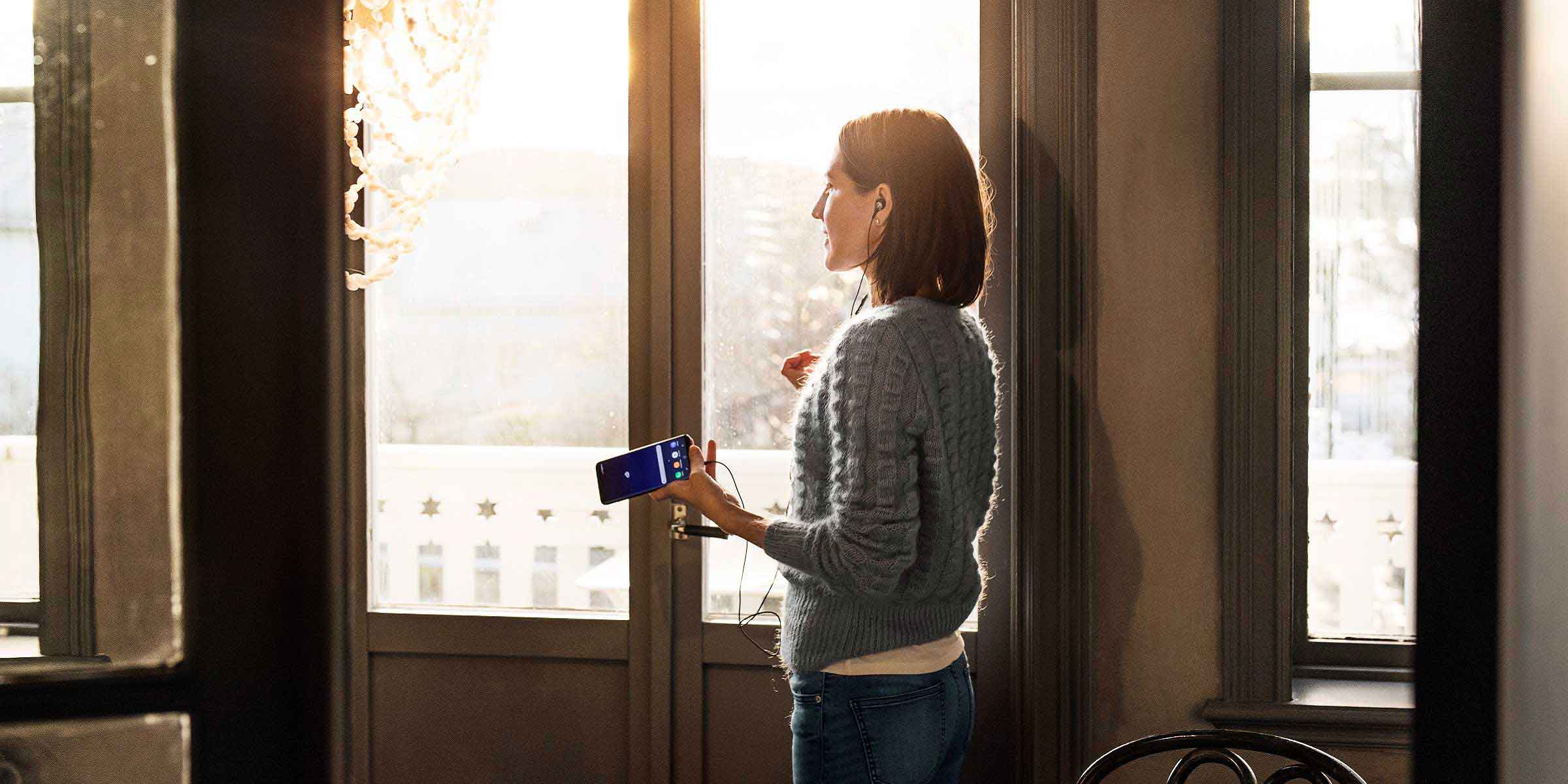 Nainen seisoo aurinkoisen ikkunan edessä puhelin kädessä.
