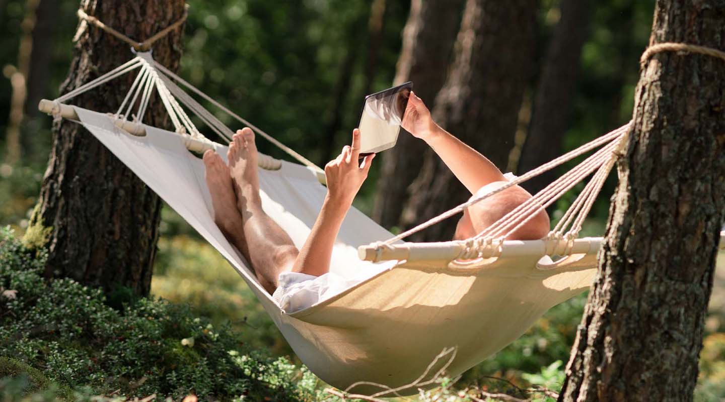 Mies makoilee riippumatossa metsässä ja katsoo tablettia