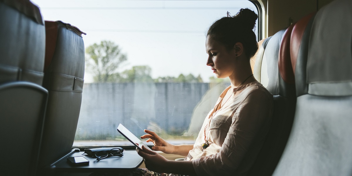 Nutturapäinen nainen istuu junassa ja käyttää tablettia.