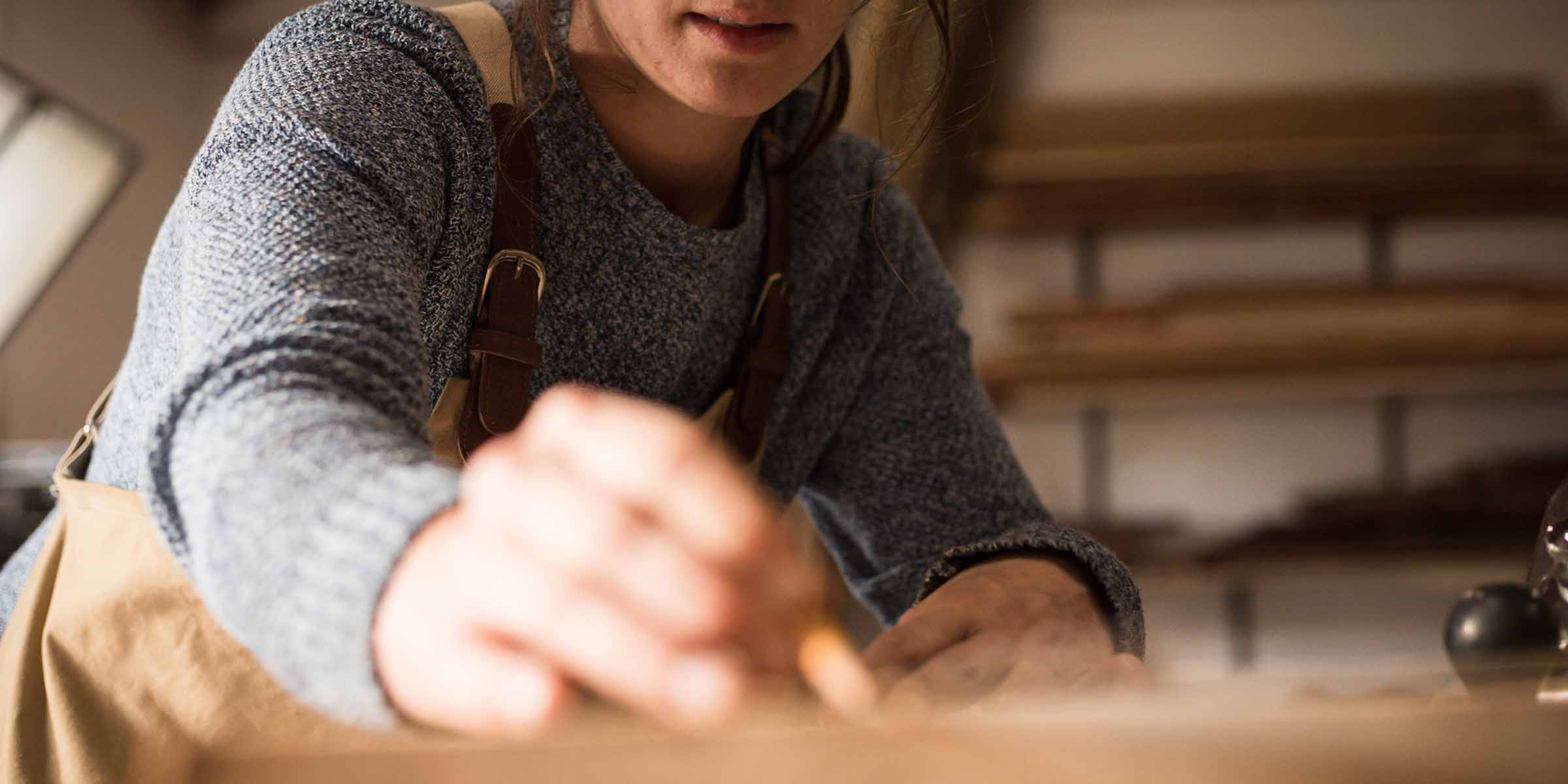 Etualalla nuoren puusepän käsi, jossa on kynä. Henkilö on kumartuneena piirustuspöydän äärelle.. 