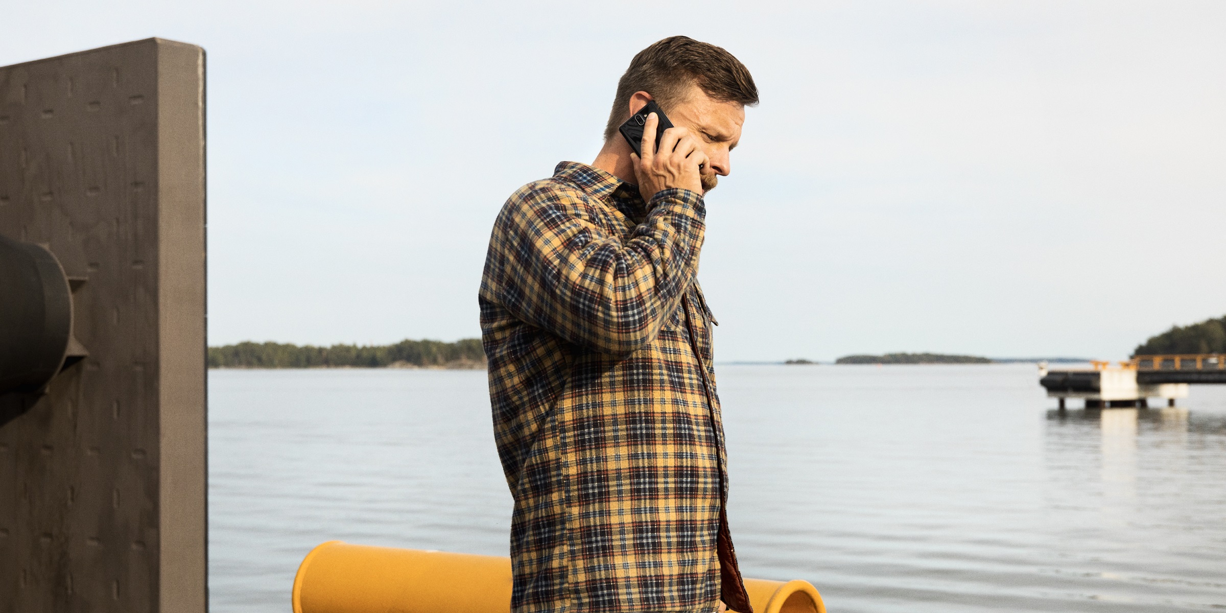 Ruutupaitainen mies laiturilla, puhuu kännykkään, taustalla järvi.
