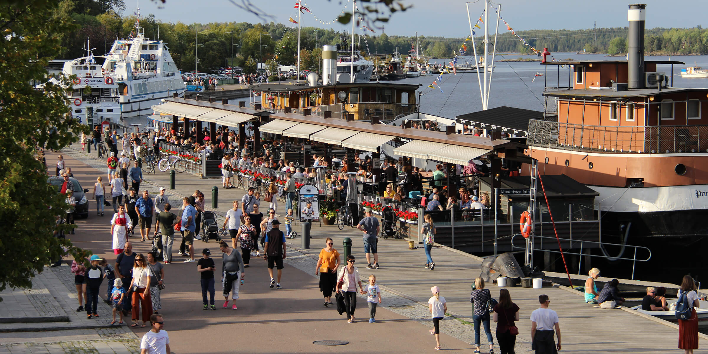 Iso joukko ihmisiä kävelee Lappeenrannan kesäisessä satamassa. Osa istuu ravintolalaivojen kyydissä. 